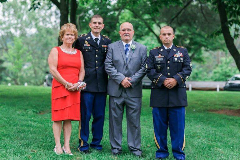 Amy, her husband, and two sons dressed in U.S. Army uniforms pose for a photo outdoors beneath lush foliage.